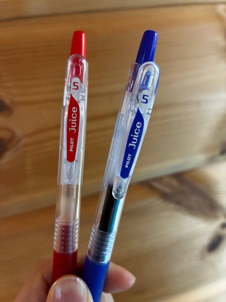A hand holding two Pilot Juice gel pens, one red and one blue, with 0.5mm tips. The pens have transparent bodies and are shown against a wooden background.