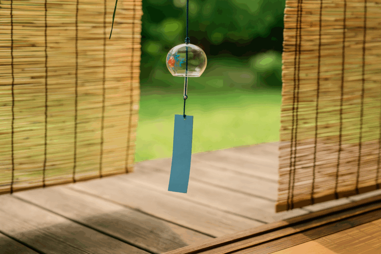 A photograph captures traditional Japanese summer items on a wooden veranda, featuring a glass wind chime, bamboo blinds, and a reed screen against a green garden backdrop.